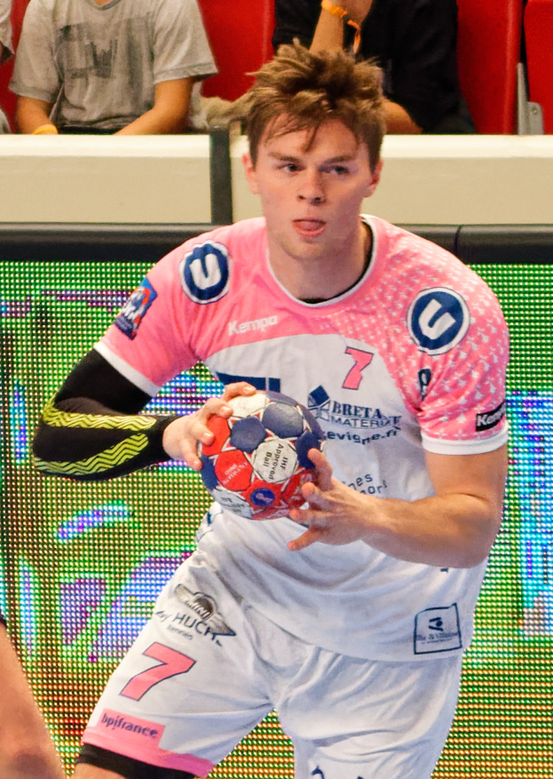 Ein Mann in einem pinken Hemd mit einem Fußball, daneben eine weitere Person und im Hintergrund Menschen auf Stühlen sitzend.