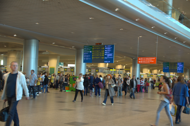 Menschen gehen durch einen Flughafen mit Hinweistafeln, Säulen, Deckenlampen und einer Glasbrüstung auf der rechten Seite.