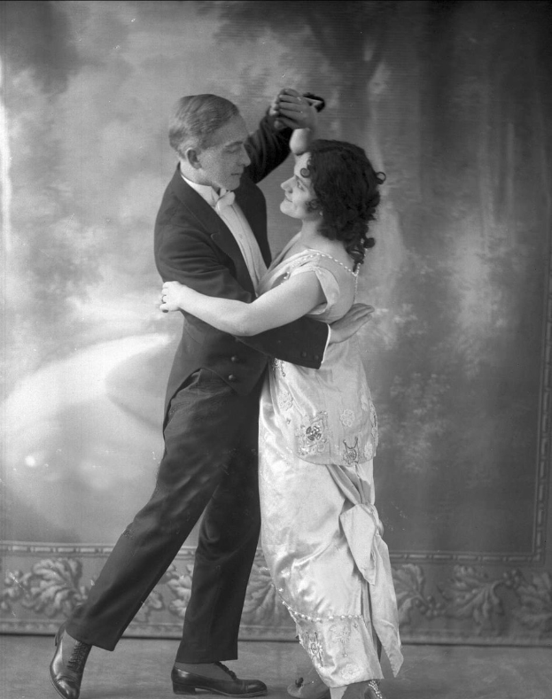 Schwarzes und weißes Foto eines Mannes in einem Anzug und einer Frau, die Tango tanzen und sich an den Händen halten, mit kunstvollen Wanddesigns im Hintergrund.