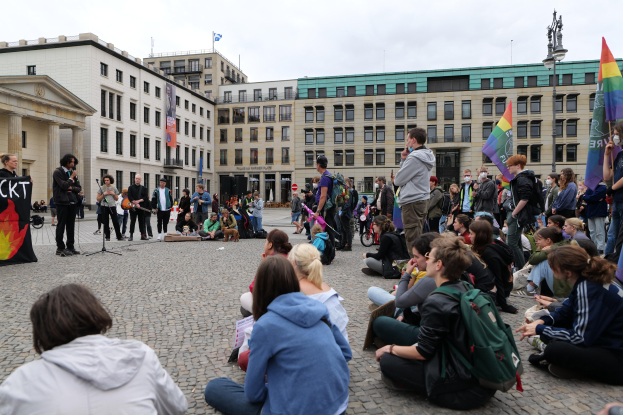 Eine Gruppe von Menschen, die auf dem Boden vor einer Menge sitzt, die Fahnen und Transparente hölt, während einer Anti-Schwulen-Demo in Berlin, mit einem Mikrofonständler, einer Statue und Gebäuden im Hintergrund.