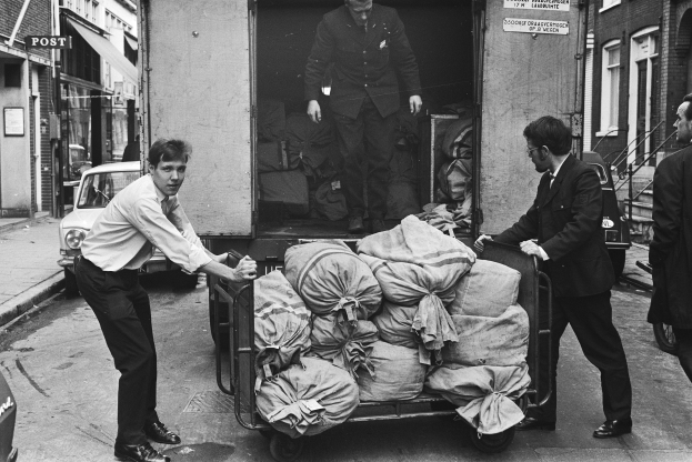 Eine Gruppe von Männern entlädt Taschen von einem geparkten Lastwagen am Straßenrand, mit einem vorbeifahrenden Auto und Gebäuden mit Fenstern, Treppen mit Geländern und Texttafeln im Hintergrund, alles in Schwarz-Weiß dargestellt.