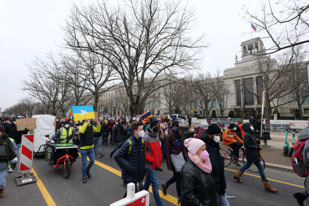 Eine große Gruppe von Menschen marschiert in Washington, D.C. demonstrierend die Straße entlang, mit Schildern und Bannern, einige auf Fahrrädern, unter einem klaren blauen Himmel.