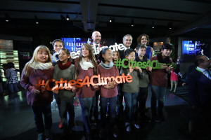 Eine Gruppe strahlender Menschen, die vor einem "Cities4Climate"-Schild zusammenstehen, mit einem hektischen Hintergrund aus Menschen, Bildschirmen und Lichtern.