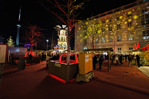 Ein lebendiges Weihnachtsmarkt in Berlin, Deutschland, mit Menschen um beleuchtete Stände, Bäume, Gebäude, Laternenmäste und einen Turm unter einem dunklen Himmel.