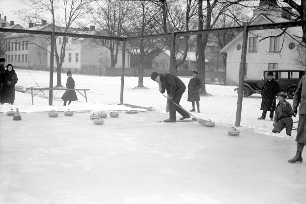 Schwarzes Bild eines Curling-Spiels auf einer Outdoor-Eisbahn, umgeben von einem Zaun, mit einer Bank in der Nähe, Bäumen, Gebäuden und einem geparkten Fahrzeug im Hintergrund.