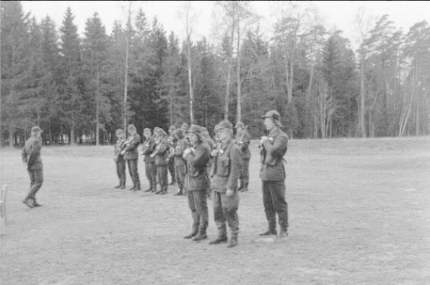 Schwarzes und weißes Bild einer Gruppe von Männern in Militäruniformen mit Mützen und Gewehren, die in einem Feld mit Bäumen und einem klaren Himmel im Hintergrund stehen.