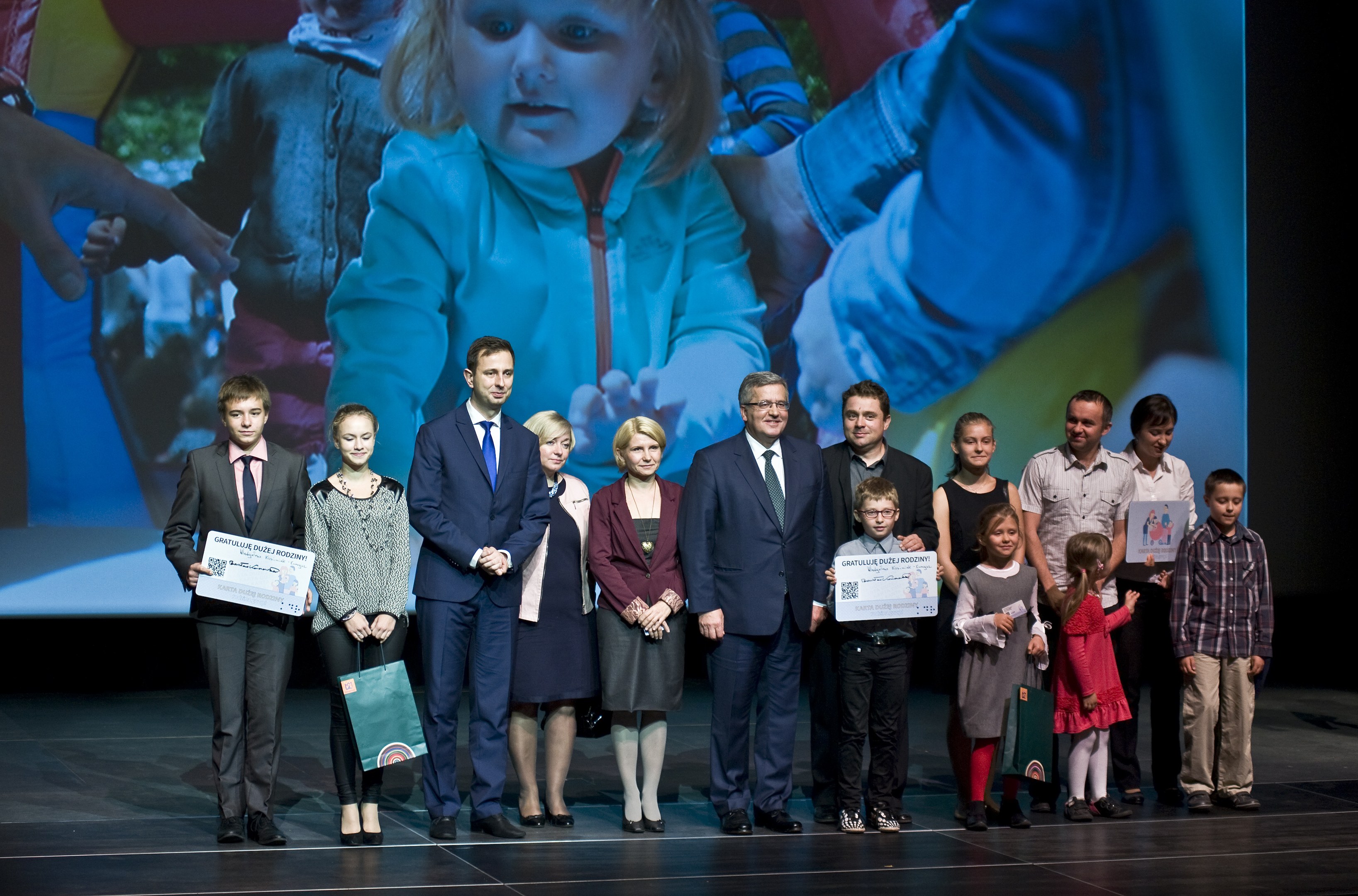 Eine Gruppe von Menschen auf einer Bühne mit Taschen und Urkunden, im Hintergrund ein Bildschirm mit einem Bild eines Kindes und anderen