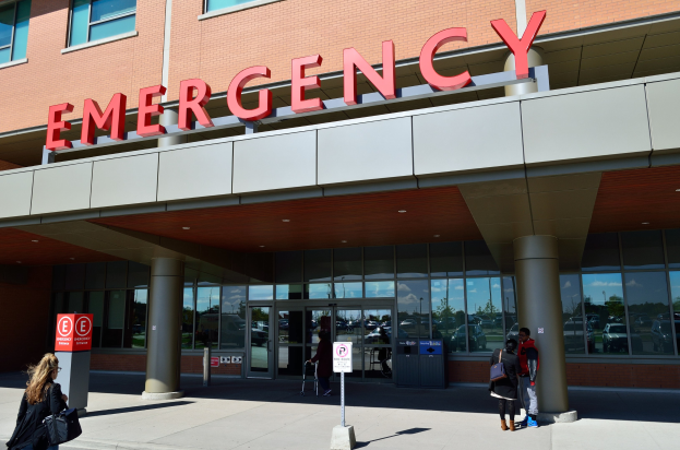 Eine Gruppe von Menschen geht vor einem Glaswand-Notfallgebäude mit Säulen, einem Namensschild, Hinweistafeln an Pfählen und einem Mülleimer vorbei.