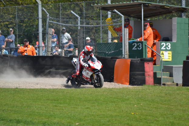 Ein Motorradfahrer mit Helm und Handschuhen fährt über ein grasbewachsenes Feld, mit Zuschauern, Absperrungen, Treppen und einem nummerierten Schuppen im Hintergrund.