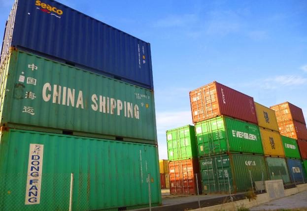 Lagercontainer in Reihen gestapelt, umgeben von einem Zaun, Gras, Pflanzen und einem bewölkten Himmel.