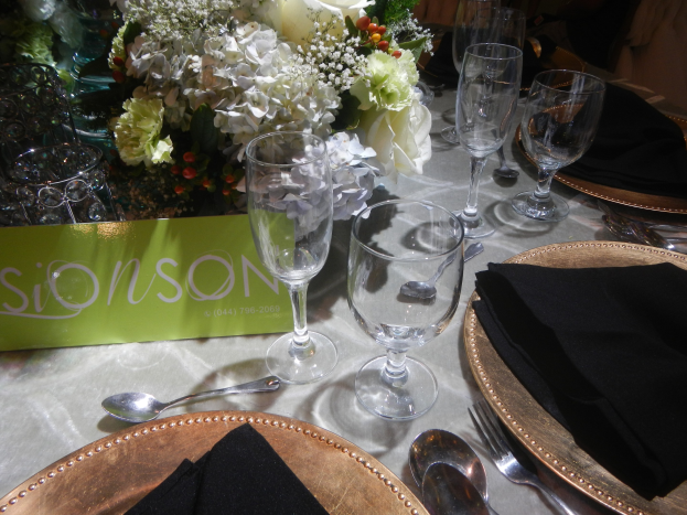 Formaler Esstisch mit goldenen Untersetzern, schwarzen Servietten, Besteck, Gläsern und einem Blumenstrauß, begleitet von einer Texttafel.