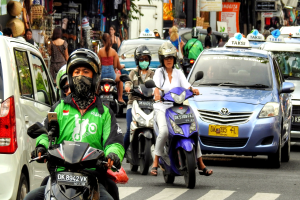 Eine Gruppe von Menschen auf Motorrädern auf einer belebten Straße mit Autos und anderen Fahrzeugen, einige Fahrer tragen Helme, Gebäude, Bäume, Schilder mit Text und Geschäfte im Hintergrund.