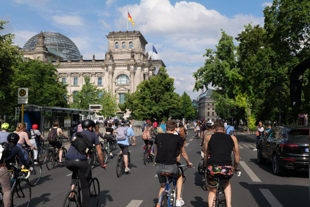 Gruppe von Menschen, die Fahrräder auf einer von Bäumen gesäumten Straße in Berlin fahren, mit einer Bushaltestelle auf der rechten Seite und einer Flagge, die auf einem Gebäude weht.