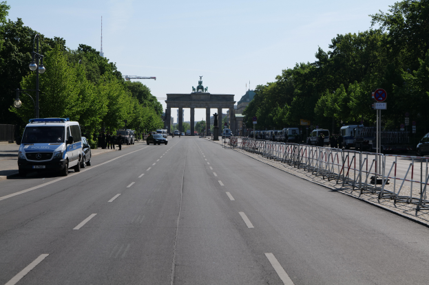 Ein Polizeiwagen steht auf der Seite einer vielbefahrenen Straße vor dem Brandenburger Tor in Berlin, Deutschland, mit Barrieren, Schildern, Bäumen, Laternenpfählen und einer bewölkten Himmel.