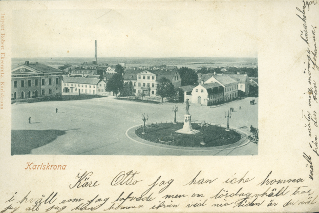 Alte Ansichtskarte zeigt eine Stadtansicht mit Gebäuden, Bäumen, Pfählen und einem zentralen Brunnen, mit handgeschriebener Text entlang der unteren Kante.