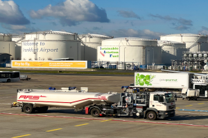 Ein weißer und grüner Tanklastwagen vor einem großen Gebäude am Frankfurter Flughafen geparkt, umgeben von anderen Fahrzeugen, mit großen weißen Tanks und einem gelben Schild im Hintergrund unter einem klaren blauen Himmel.