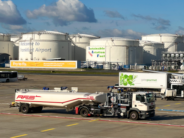 Ein weißer und grüner Tanklastwagen vor einem großen Gebäude am Frankfurter Flughafen geparkt, umgeben von anderen Fahrzeugen, mit großen weißen Tanks und einem gelben Schild im Hintergrund unter einem klaren blauen Himmel.