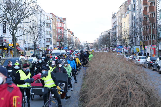Eine große Gruppe von Menschen mit Masken und Sicherheitswesten auf Fahrrädern auf einer von Bäumen gesäumten Straße mit Gebäuden, Fahrzeugen, Laternenmästen und Texttafeln unter einem klaren blauen Himmel.