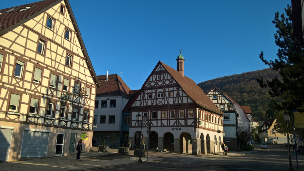 Eine kleine Stadt mit Gebäuden, Straßenlaternen, Fahrzeugen, Fußgängern, Infotafeln, Bäumen und Hügeln unter einem klaren blauen Himmel, mit einem zentralen Haus.