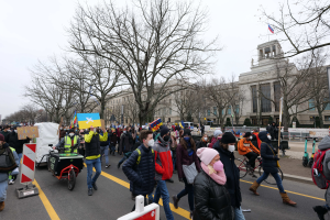 Eine große Gruppe von Menschen nimmt an einer Protestdemo auf einer Straße in Washington, D.C. teil, einige halten Schilder und Banner, andere fahren Fahrräder, und Schilder, Bäume und ein klarer blauer Himmel sind im Hintergrund zu sehen.