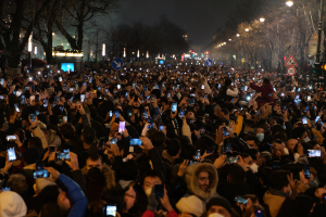 Eine große Menschenmenge, die nachts auf einer Straße steht, beleuchtet von Straßenlaternen, umgeben von Bäumen und Schildern, viele halten ihre Mobiltelefone hoch.