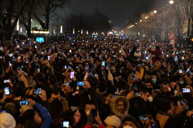 Eine große Menschenmenge, die nachts auf einer Straße steht, beleuchtet von Straßenlaternen, umgeben von Bäumen und Schildern, viele halten ihre Mobiltelefone hoch.