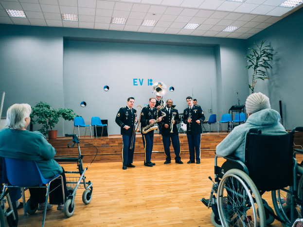 Eine Gruppe von Menschen im Rollstuhl, die in einem Raum mit Stühlen, Pflanzen und Deckenleuchten Musikinstrumente spielen, während einige Menschen auf Stühlen sitzen.