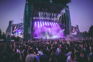 Eine große Menschenmenge versammelt sich auf einem Musikfestival, mit einer Bühne in der Mitte, auf der Musiker Instrumente spielen, beleuchtet von Scheinwerfern und umgeben von Bildschirmen, vor dem Hintergrund eines Gebäudes, Bäume und eines klaren blauen Himmels.