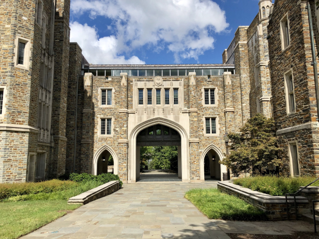 Großes steinernes Universitätsgebäude mit gewölbtem Eingang, umgeben von grünem Rasen, Bäumen und weißen Wolken am Himmel.