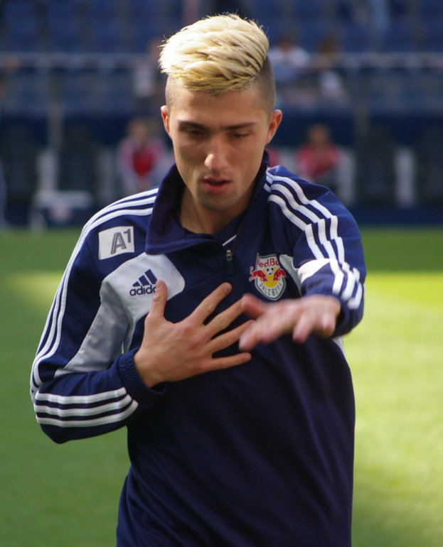 Ein junger Mann in einem blauen T-Shirt steht auf einem Fußballfeld und streckt die Arme aus.