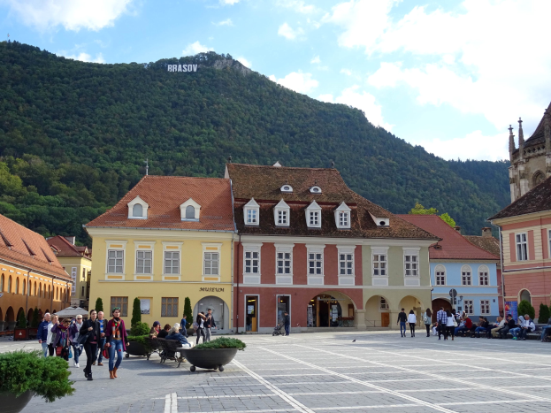 Menschen, die in einem Stadtplatz mit Gebäuden, Fenstern, Laternen und Topfpflanzen umhergehen, mit einem Hügel und einem bewölkten Himmel im Hintergrund.