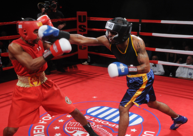 Zwei Boxer kämpfen in einem Ring, während ein Mann im Hintergrund filmt, mit einer kleinen Zuschauermenge im dunklen Hintergrund.