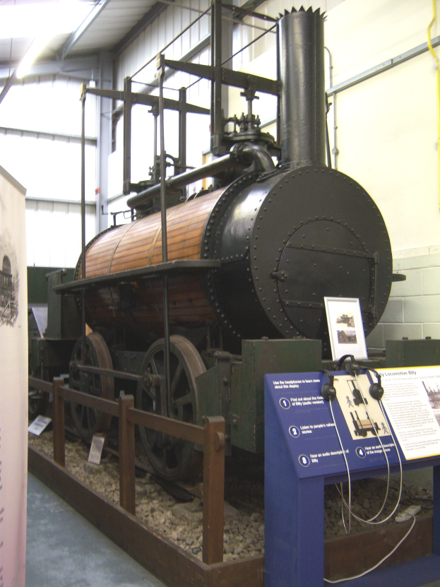 Alte Dampflokomotive in einem Museum mit Informationsschildern, Zaun und umgebenen Ausstellungen.