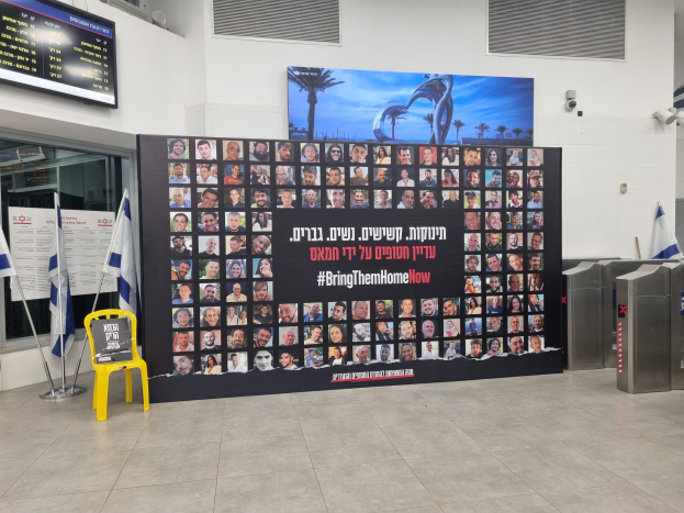 Eine große Wand zeigt verschiedene Bilder, einen gelben Stuhl, israelische Regierungsflaggen auf Stangen, eine Tafel mit der Aufschrift "Bring Them Home Now", einen montierten Bildschirm, Deckenleuchten und andere Gegenstände.