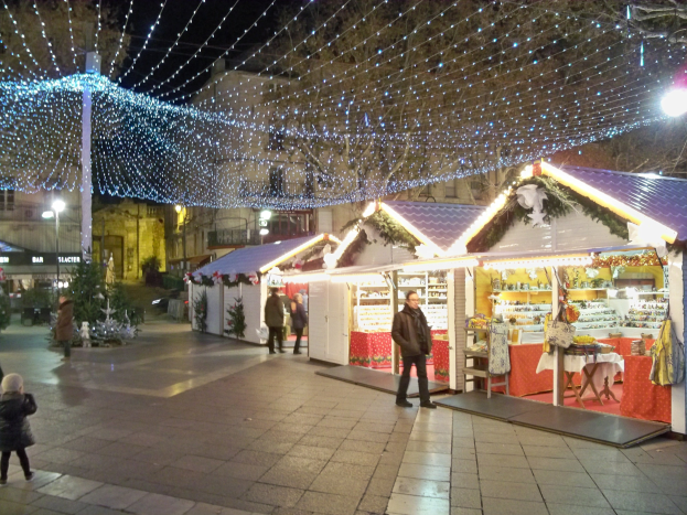 Ein geschäftiger Weihnachtsmarkt bei Nacht in einer Stadt, mit Menschen, die stehen, sitzen und ein Kind halten, dekoriert mit festlichen Lichtern, Pflanzen, Bäumen und Gebäuden unter einem bewölkten Himmel.