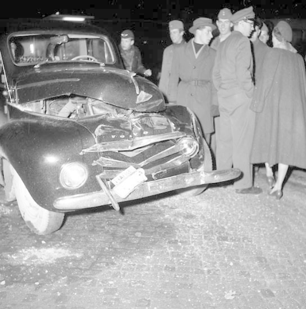 Schwarz-weißes Bild eines schwer beschädigten Autos am Straßenrand mit einer Gruppe von Menschen, die es untersuchen.