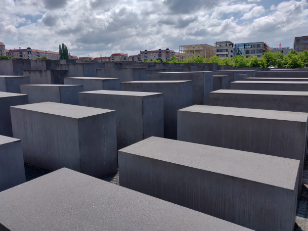 Konkreter Holocaust-Denkmal mit rechteckigen Blöcken, die in einem Gittermuster angeordnet sind und von Bäumen, Gebäuden und einem bewölkten Himmel umgeben sind.