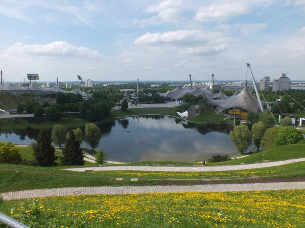 Luftaufnahme des Olympiastadions von einem Hügel aus, mit einem Teich im Vordergrund, umgeben von Grün und gelben Blumen, und im Hintergrund sichtbare Geb├Ąude unter einem klaren blauen Himmel.