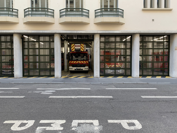 Feuerwache mit Feuerwehrauto davor, mit Fenstern, Geländern, Säulen und Glastüren, mit einer Straße unten.