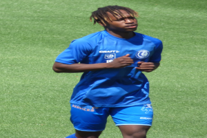 Ein junger Mann in einem blauen T-Shirt und Shorts, der auf einem Fußballfeld läuft und die Arme ausstreckt.