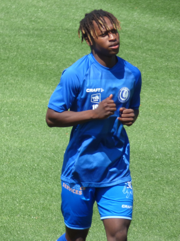 Ein junger Mann in einem blauen T-Shirt und Shorts, der auf einem Fußballfeld läuft und die Arme ausstreckt.