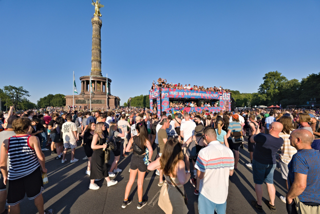 Große Menschenmenge versammelt sich vor einem Berliner Denkmal mit Fahnen, einer säulengeschmückten Gebäude, Statue, Inschrift, Bäumen und wolkenlosem Himmel.