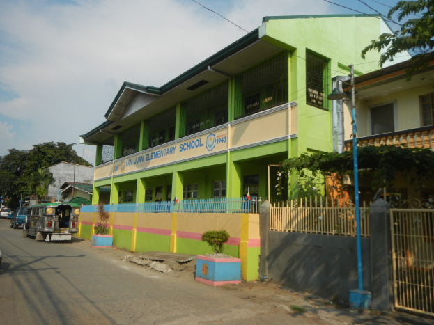 Schulgebäude mit grüner und gelber Fassade, Fahrzeugen auf der Straße, Topfpflanzen, einem Zaun, einem Tor, einem Straßenpfahl, Bäumen, Drähten und einem bewölkten Himmel.