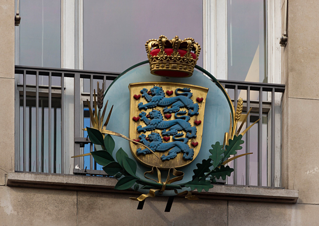 Wappen der britischen Königsfamilie an einer Gebäudewand mit einer Krone- und Blatt-Logo, Fenstern und Metallgittern im Hintergrund.