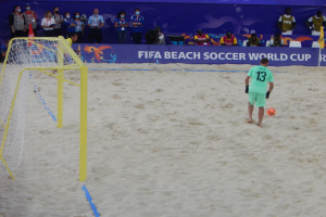 Ein Mann schießt einen Fußball auf einem sandigen Strand während eines Spiels, mit einem Tor auf der linken Seite und Zuschauern, einige halten Kameras, im Hintergrund.