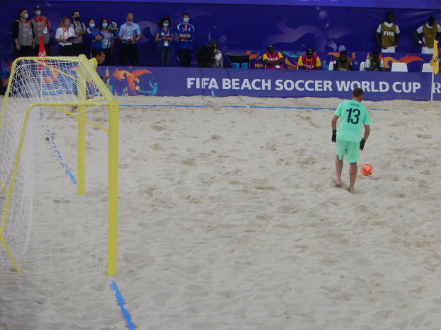Ein Mann schießt einen Fußball auf einem sandigen Strand während eines Spiels, mit einem Tor auf der linken Seite und Zuschauern, einige halten Kameras, im Hintergrund.