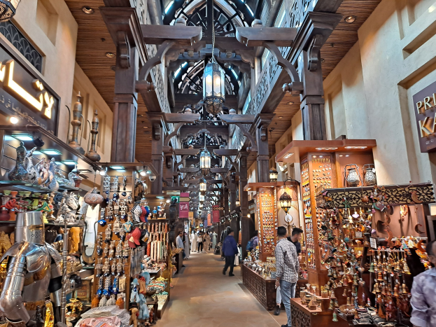 Ein belebter Dubai-Basar mit Menschen, die zwischen Regalen voller Waren gehen, beleuchtet von Deckenlampen und Wänden mit Texttafeln.