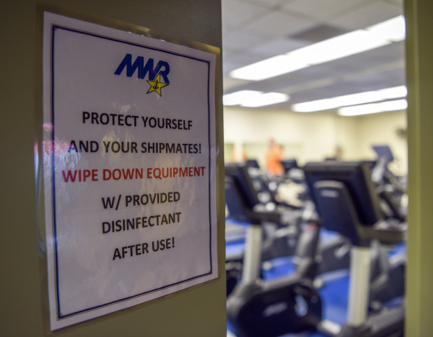 Zeichen an einer Turnhalle mit der Aufschrift "Schützen Sie sich und Ihre Schiffskameraden" mit Laufbändern und Deckenleuchten im Hintergrund.