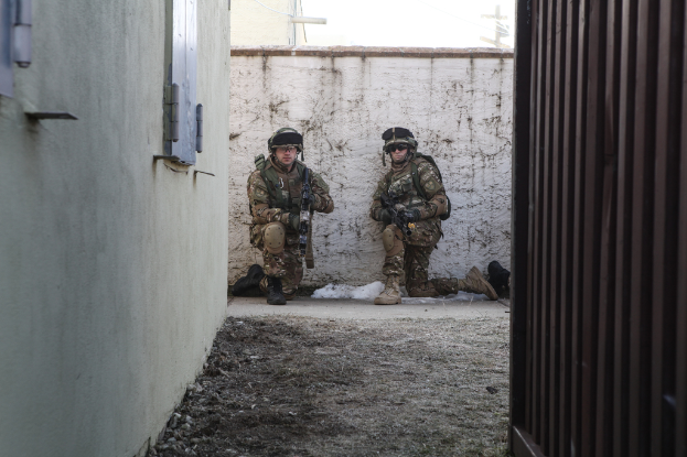 Eine Szene mit einer Wand und einem Fenster auf der linken Seite, einer eisernen Wand auf der rechten Seite, zwei Soldaten und Erde in der Mitte und Gebäuden, einem Strommast und einer weiteren Wand im Hintergrund.
