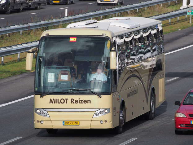Ein Minibus mit sichtbaren Insassen auf einer Autobahn neben einem roten Auto fahrend, mit Geländern und Gras am Straßenrand und Textanzeigetafeln und Säulen im Hintergrund.